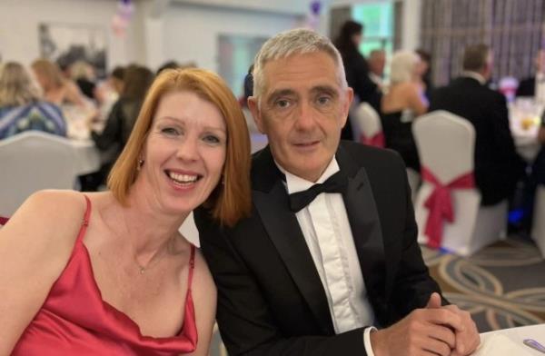 Toni with Paul wearing black tie at an event, sitting on their table and smiling