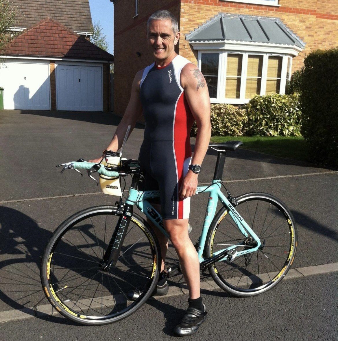 Paul posing with his bike outside his house, smiling and wearing a leotard